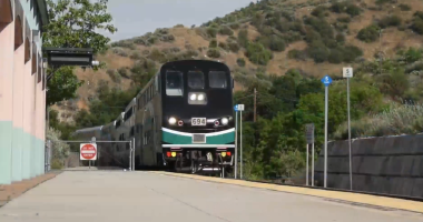A Metrolink commuter train approaches a station platform in a hilly area, with the train number 694 visible on the front and signs indicating car stops along the platform.